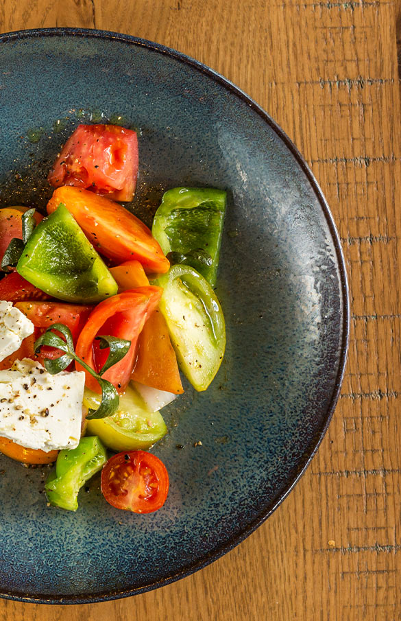 Greek Salad in bowl with large pieces of heirloom tomatoes, marinated feta, red onion, bell pepper in cucumber vinaigrette.