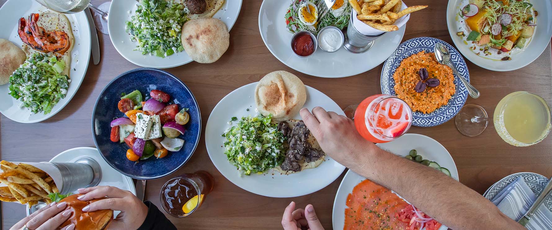 A large spread of mediterranean food from Del Mar Naples sits on a wooden table.